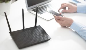 Router on desk with multiple antennas, laptop in background, person using smartphone, illustrating network hardware and custom firmware implications for information technology education.