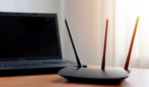 Router with three antennas on a wooden desk next to a laptop, illustrating the hardware used in networking and the importance of router firmware in information security education.