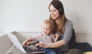 Young Caucasian mother student with baby working on laptop from home. Workplace of freelance woman student with kid toddler. 