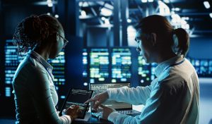 Engineering colleagues running code on tablet and laptop, updating professional data center equipment designed to accommodate server racks, networking gadgets and storage arrays