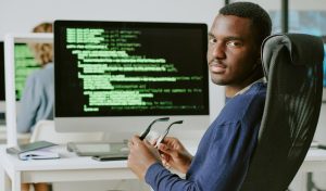Young African American male cybersecurity specialist sitting at workplace taking off his eyeglasses and looking at camera