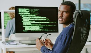Young African American male cybersecurity specialist sitting at workplace taking off his eyeglasses and looking at camera