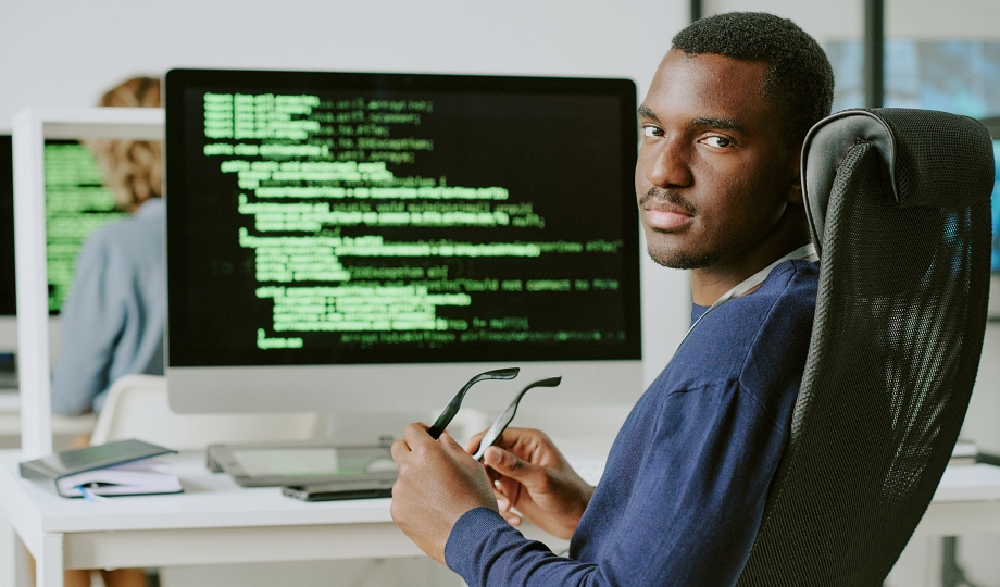 Young African American male cybersecurity specialist sitting at workplace taking off his eyeglasses and looking at camera