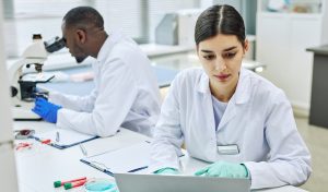 Lab technicians examining samples under a microscope and reviewing data on a laptop