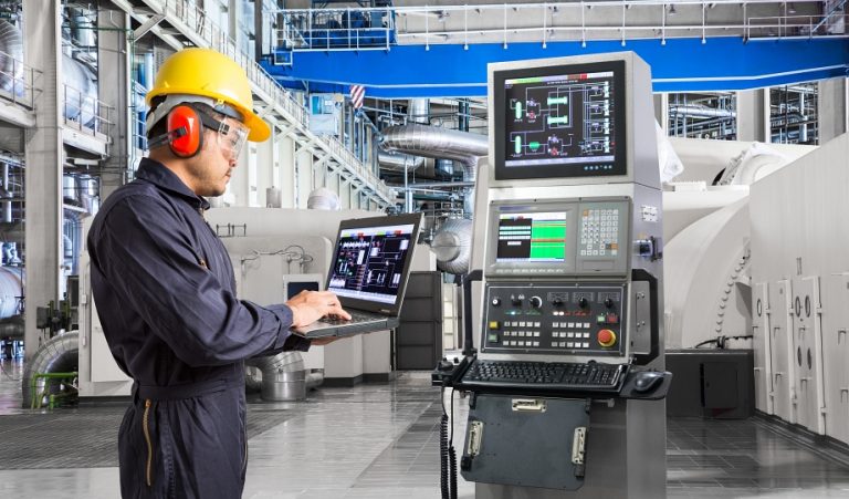 Instrument calibration technician in a yellow hard hat and ear protection using a laptop at an industrial control station