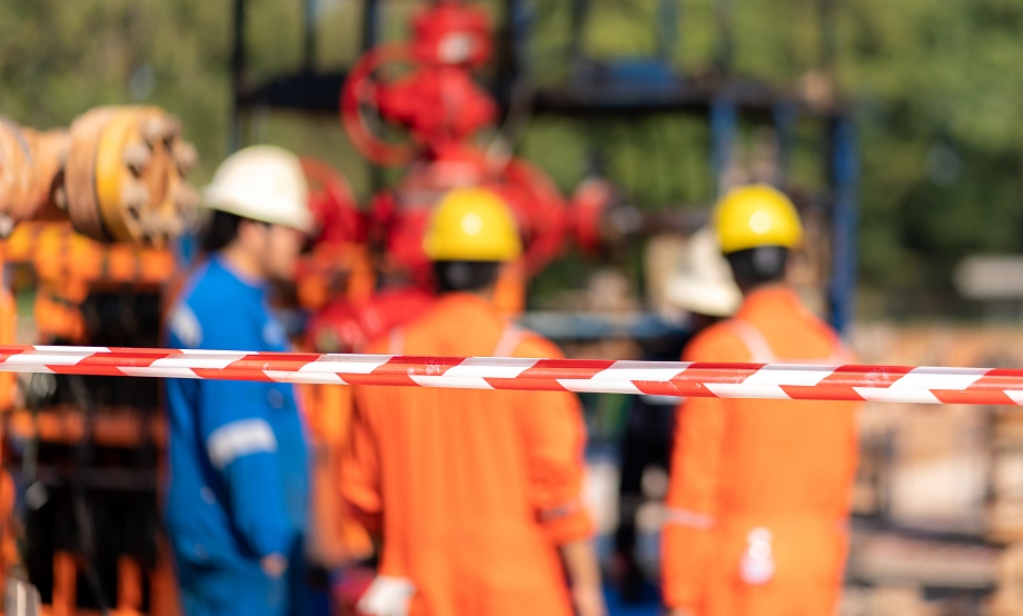 Plant workers in hard hats and orange coveralls behind red-and-white caution tape at an industrial site