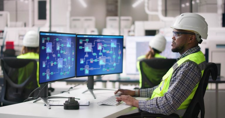 Process technician in a white hard hat and safety vest monitoring process diagrams on two computer screens