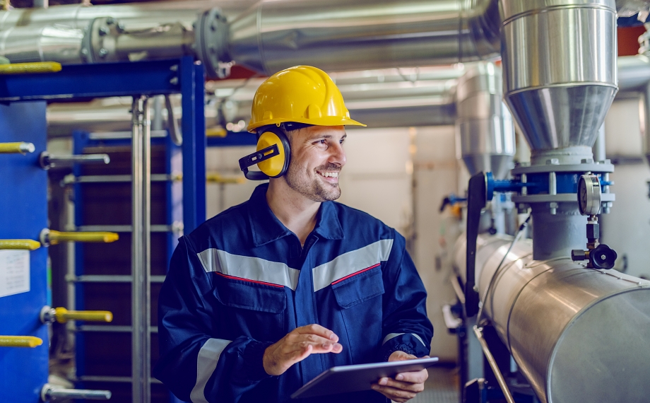Smiling process technician in a yellow hard hat and ear protection holding a tablet next to plant piping
