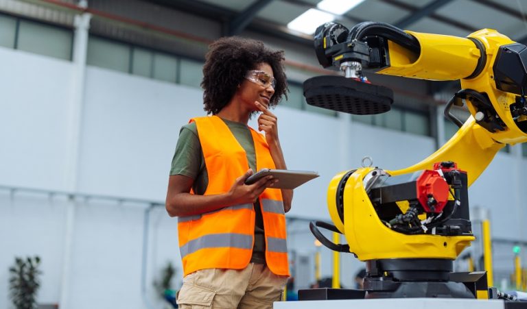 Woman holding a tablet beside a yellow industrial robot arm in a factory that uses SCADA
