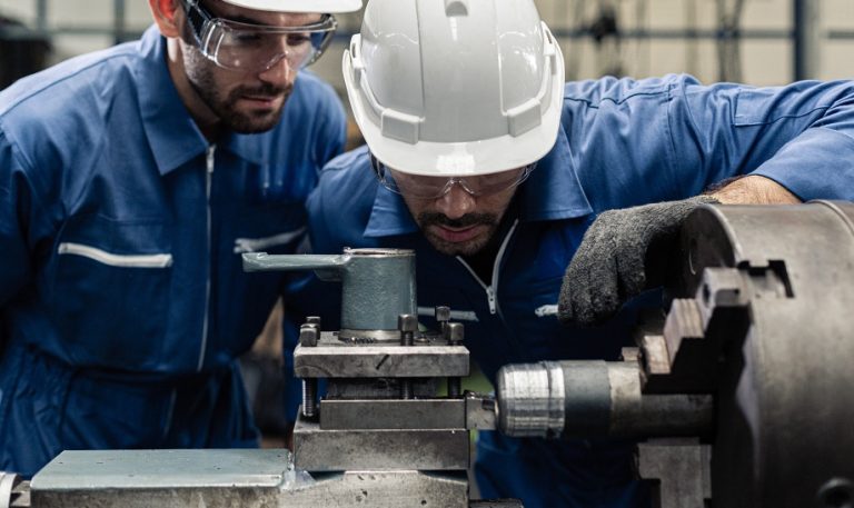 Two technicians in blue coveralls and safety glasses inspecting a machine while troubleshooting a system failure